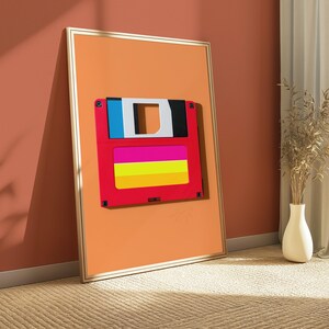 May include: A framed vertical wall art print featuring a stylised graphic of a 3.5-inch floppy disk. The disk is red with a blue and white top section and a horizontal label area containing bright pink, yellow, and orange stripes on an orange background.