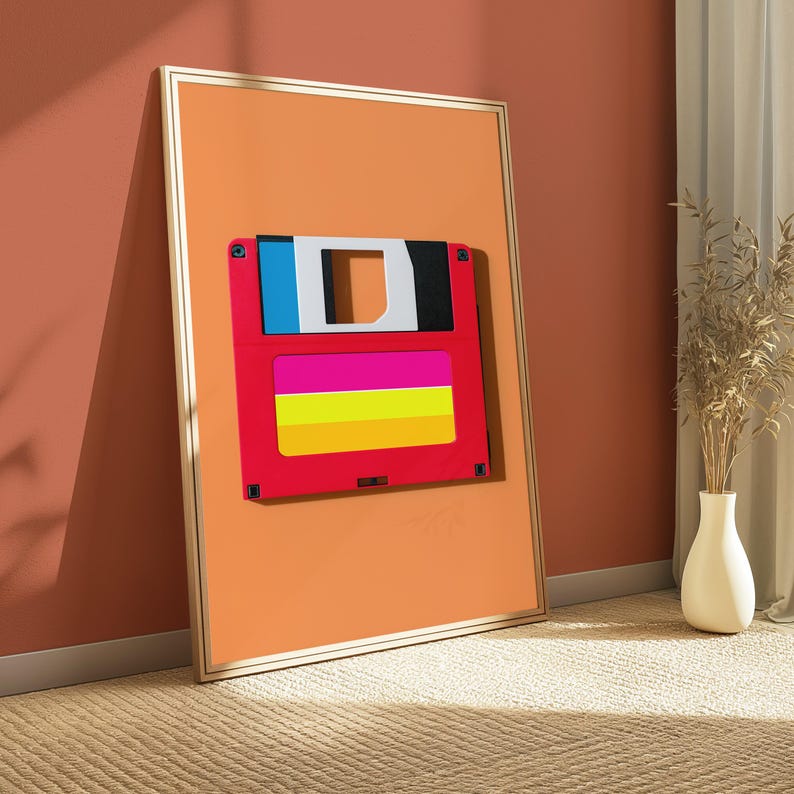 May include: A framed vertical wall art print featuring a stylised graphic of a 3.5-inch floppy disk. The disk is red with a blue and white top section and a horizontal label area containing bright pink, yellow, and orange stripes on an orange background.