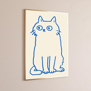 May include: A framed wall art print featuring a simple, hand-drawn style illustration of a cat in vibrant blue ink. The cat is depicted with wide, circular eyes and a small nose, set against a solid, light cream-colored background.