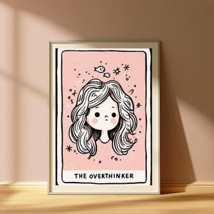 May include: A framed art print featuring a pale pink background with a black-outlined illustration of a person with wavy hair, rosy cheeks, and small fish and star icons floating above. The text THE OVERTHINKER is printed in black at the bottom.
