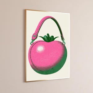 May include: A framed wall art print featuring a stylized illustration of a pink tomato-shaped handbag with a green stem top. The bag has a pink strap with green edges, dark green shading, and visible stitching details in a halftone dot pattern.