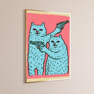 May include: A framed wall art print featuring two light blue cats with red eyes against a solid pink background. Both cats are depicted in a cartoon style, each holding a black revolver. The artwork is enclosed in a thin gold-coloured frame.