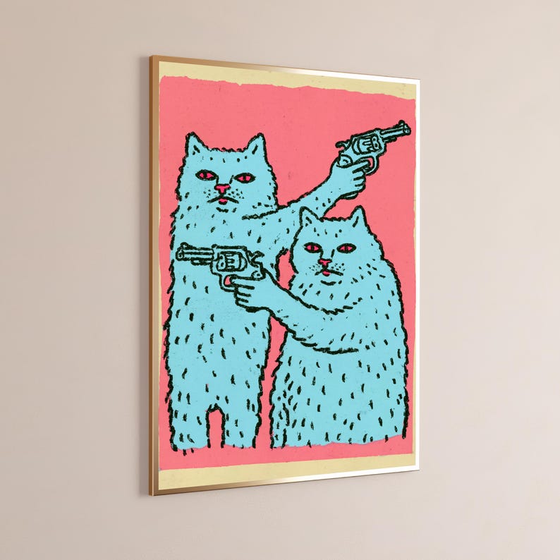 May include: A framed wall art print featuring two light blue cats with red eyes against a solid pink background. Both cats are depicted in a cartoon style, each holding a black revolver. The artwork is enclosed in a thin gold-coloured frame.