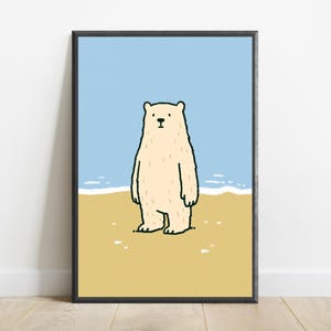May include: A framed wall art print featuring a cartoon illustration of a cream-colored polar bear standing on a sandy yellow beach. The background shows a light blue sky and a thin white line representing the ocean surf behind the bear.
