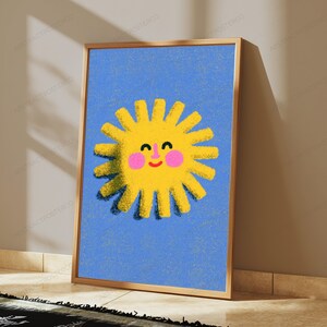 May include: A framed wall art print featuring a bright yellow sun with a smiling face, pink cheeks, and a textured appearance centred on a solid blue background. The sun has numerous radiating rays and a dark shadow effect on its lower left side.