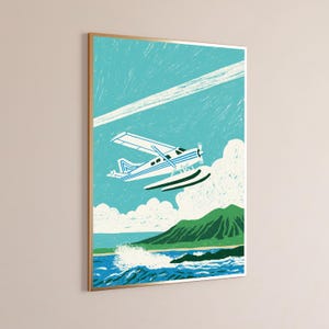 May include: A framed art print featuring a white and blue seaplane flying over a green mountain and blue ocean waves. The sky is a textured turquoise with a white contrail and fluffy white clouds above a sandy beach and dark green coastline.