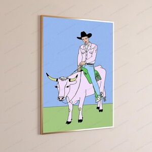 May include: A framed wall art print featuring a line drawing of a person in a black cowboy hat, pink jacket, and blue jeans riding a light pink bull. The bull has yellow horns and stands on a green field against a light blue sky background.