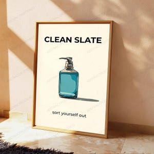 May include: A framed wall art print featuring a cream-colored background with a central illustration of a blue liquid soap dispenser. The text CLEAN SLATE is printed in black above the dispenser, and the text sort yourself out is below it.