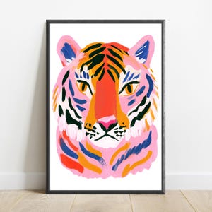 May include: A framed art print featuring a stylized tiger head illustration. The tiger is rendered in vibrant shades of pink, orange, and blue, with bold black stripes and piercing yellow eyes, all set against a clean white background.