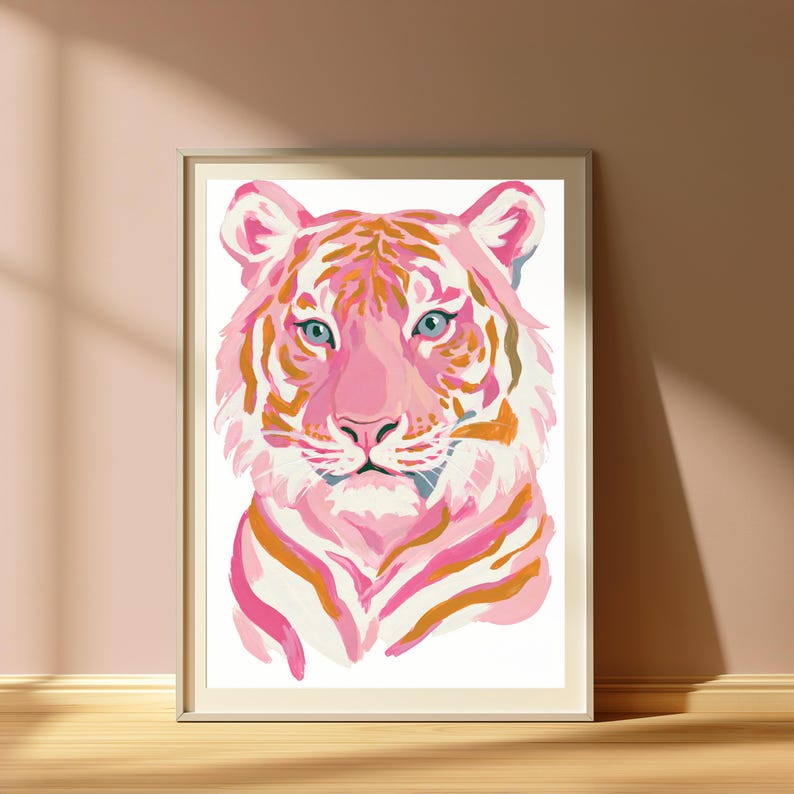May include: A framed wall art print featuring a stylized tiger portrait. The tiger is rendered in shades of light pink, magenta, and warm orange, with distinct stripes and piercing blue eyes. The artwork is set against a clean white background.