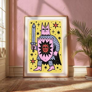 May include: A framed art print featuring a stylised illustration of a horned figure in pink armour holding a grey sword and a pink shield with a red sun design. The background is yellow with black stars and crosses, all enclosed in a gold frame.
