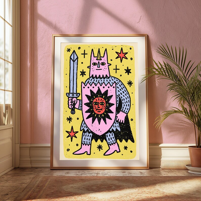 May include: A framed art print featuring a stylised illustration of a horned figure in pink armour holding a grey sword and a pink shield with a red sun design. The background is yellow with black stars and crosses, all enclosed in a gold frame.