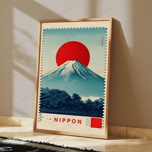 K&ouml;nnte beinhalten: Ein gerahmter, rechteckiger Wandkunstdruck, der wie eine Briefmarke gestaltet ist. Das Bild zeigt eine gro&szlig;e rote Sonne &uuml;ber dem blau get&ouml;nten Berg Fuji, mit dunklen B&auml;umen im Vordergrund und dem Schriftzug NIPPON am unteren Rand.