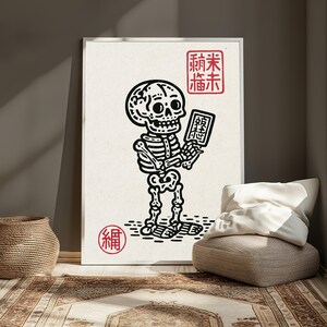 May include: A framed wall art print featuring a black ink illustration of a skeleton holding a small rectangular tablet. The design includes red stamp-like symbols with East Asian characters in the top right and bottom left corners on a cream background.