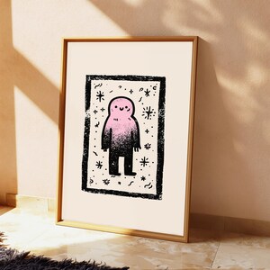 May include: A framed art print featuring a central pink and black gradient creature with a simple smiling face. The figure is enclosed within a thick, textured black rectangular border, surrounded by small black star and squiggle motifs on a cream background.