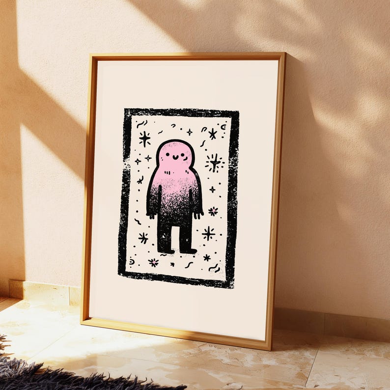 May include: A framed art print featuring a central pink and black gradient creature with a simple smiling face. The figure is enclosed within a thick, textured black rectangular border, surrounded by small black star and squiggle motifs on a cream background.