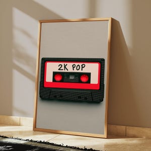 May include: A framed vertical wall art print featuring a black cassette tape with a bright red centre label and the text 2K POP written in black marker. The cassette has two red circular spools and is centred on a light beige background.