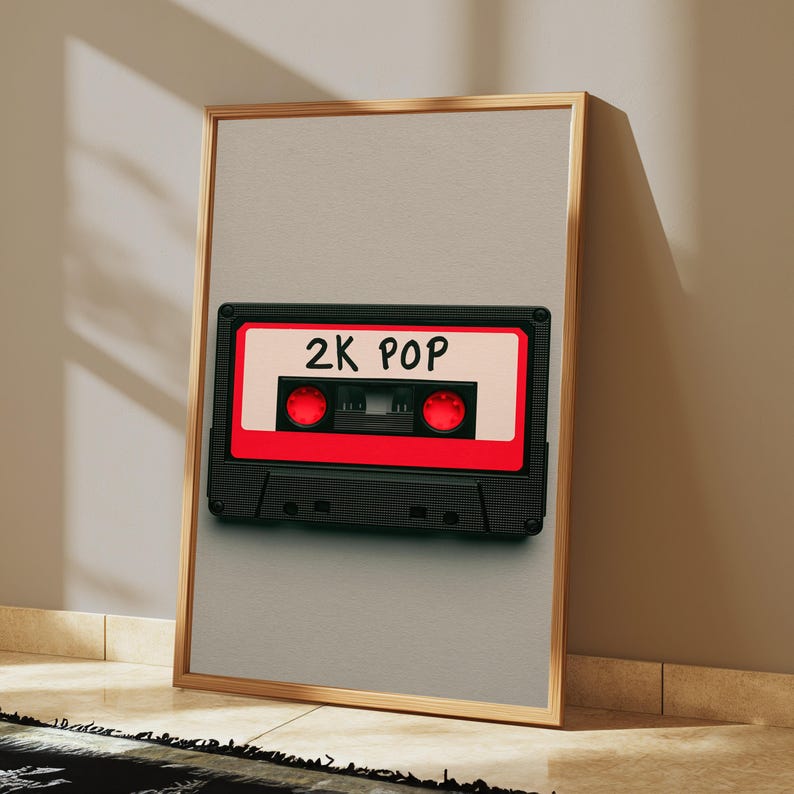May include: A framed vertical wall art print featuring a black cassette tape with a bright red centre label and the text 2K POP written in black marker. The cassette has two red circular spools and is centred on a light beige background.