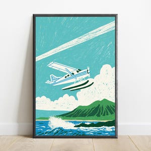 May include: A framed print featuring a white and blue seaplane flying over a turquoise ocean with white waves, a green mountain range, and white clouds against a teal sky with a diagonal white contrail line across the upper portion.