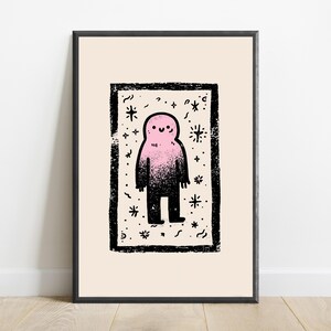 May include: A framed art print featuring a central rectangular black border containing a stylized pink and black gradient figure with a simple smiling face, surrounded by small black star and squiggle motifs on a light cream background.