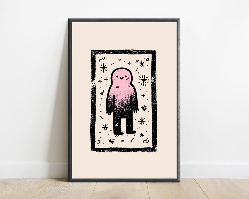 May include: A framed art print featuring a central rectangular black border containing a stylized pink and black gradient figure with a simple smiling face, surrounded by small black star and squiggle motifs on a light cream background.