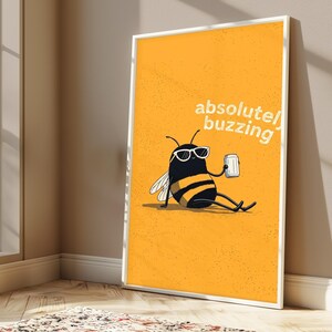 May include: A framed rectangular wall art print featuring a yellow background with a black and yellow bee wearing white sunglasses, sitting and holding a silver can. The text "absolutely buzzing" is printed in white in the upper right corner.