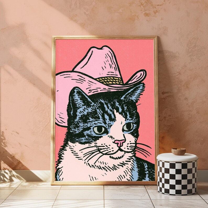 May include: A framed wall art print featuring a black and white tabby cat wearing a light pink cowboy hat. The cat is depicted in a graphic illustration style against a solid pink background, with visible black ink outlines and a light pink nose.