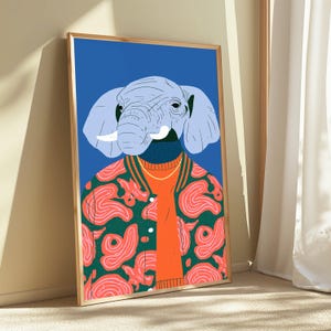 May include: A framed wall art print featuring a stylized illustration of an elephant head with light blue skin and white tusks. The elephant wears a bright orange sweater under a green varsity jacket with a pink swirling pattern on a blue background.