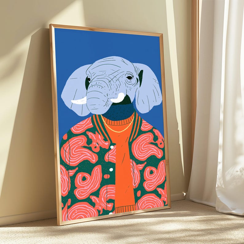 May include: A framed wall art print featuring a stylized illustration of an elephant head with light blue skin and white tusks. The elephant wears a bright orange sweater under a green varsity jacket with a pink swirling pattern on a blue background.