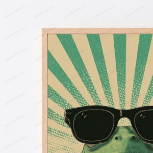 May include: A framed wall art print featuring a green frog wearing large black sunglasses. The background consists of a retro-style sunburst pattern in alternating shades of muted green and beige with a subtle speckled texture throughout the design.