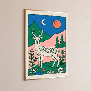 May include: A framed wall art print featuring a white deer with antlers standing in a landscape with green mountains, pink hills, and a blue night sky with a crescent moon and a round orange sun. The foreground includes various flowers and plants.