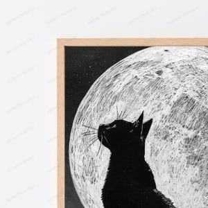 May include: A framed wall art print featuring a solid black cat silhouette in profile, looking upward toward a large, textured white full moon against a dark, starry night sky. The artwork is encased in a light-toned wooden frame.