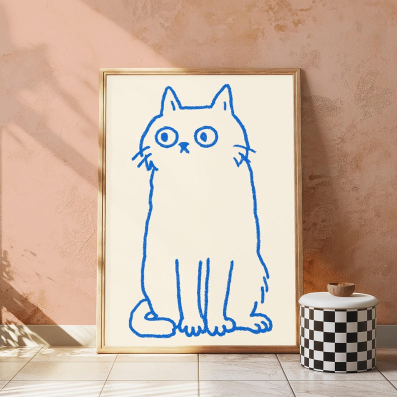 May include: A rectangular framed wall art piece featuring a simple, bold blue line drawing of a cat with large, wide eyes on a cream-colored background. The cat is depicted in a seated position with a thick, hand-drawn style outline.