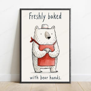 Puede incluir: Una l&aacute;mina enmarcada que muestra un oso blanco con un delantal rojo y un peque&ntilde;o gorro de chef blanco. El oso est&aacute; sobre un fondo crema con el texto "Freshly baked" en la parte superior y "with bear hands." en la parte inferior en negro.