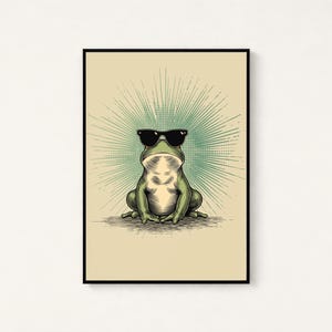 May include: A framed wall art print featuring a green frog wearing black sunglasses. The frog is centered against a light beige background with a radiating teal sunburst pattern emanating from behind its head, enclosed in a thin black frame.