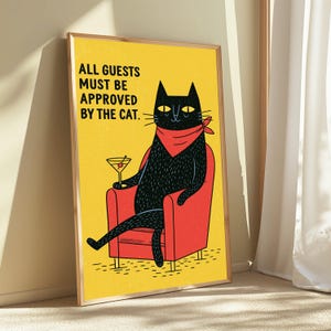 May include: A framed wall art print featuring a black cat wearing a red bandana, sitting in a red armchair while holding a martini glass. The background is yellow with the black text: ALL GUESTS MUST BE APPROVED BY THE CAT. The print is in a wooden frame.