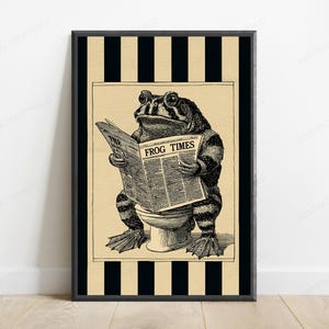May include: A framed wall art print featuring a black and white illustration of a toad sitting on a toilet reading a newspaper titled The Frog Times. The background consists of vertical black and beige stripes, framing the central toad character.