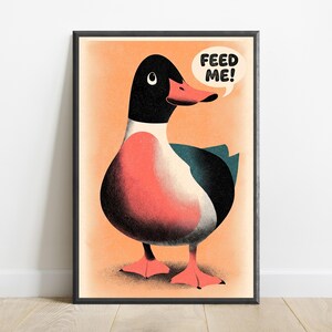 May include: A framed art print featuring a stylised illustration of a duck with a black head, a coral-coloured chest, and dark wings. The duck has a coral beak and webbed feet, set against a light orange background with a speech bubble saying FEED ME!