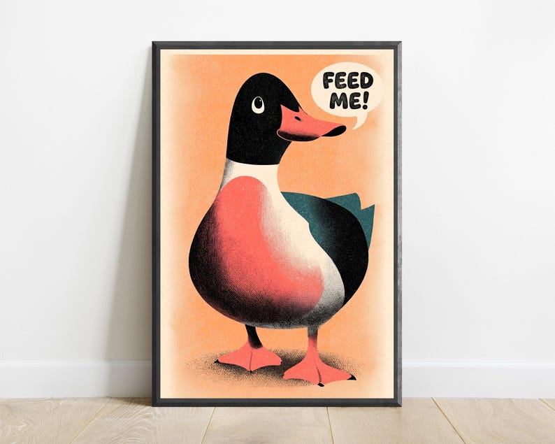 May include: A framed art print featuring a stylised illustration of a duck with a black head, a coral-coloured chest, and dark wings. The duck has a coral beak and webbed feet, set against a light orange background with a speech bubble saying FEED ME!