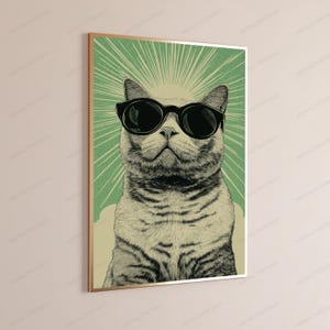 May include: A framed wall art print featuring a detailed illustration of a tabby cat wearing large black sunglasses. The cat is centered against a light green background with radiating white sunburst lines extending from behind its head.
