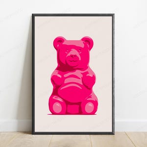 May include: A framed wall art print featuring a centered, stylized illustration of a bright pink gummy bear. The bear is depicted with a smooth, glossy appearance against a solid, light beige background, enclosed within a thin black frame.