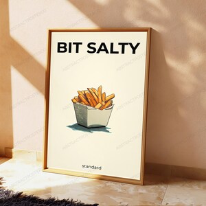 K&ouml;nnte beinhalten: Ein rechteckiges Wandbild in einem Holzrahmen mit cremefarbenem Hintergrund, dem schwarzen Text BIT SALTY oben und standard unten. In der Mitte befindet sich eine grafische Illustration von goldbraunen Pommes frites in einer wei&szlig;en Schachtel.