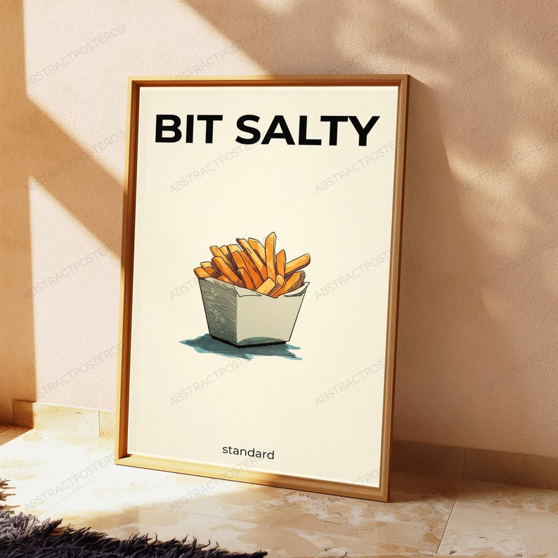 K&ouml;nnte beinhalten: Ein rechteckiges Wandbild in einem Holzrahmen mit cremefarbenem Hintergrund, dem schwarzen Text BIT SALTY oben und standard unten. In der Mitte befindet sich eine grafische Illustration von goldbraunen Pommes frites in einer wei&szlig;en Schachtel.