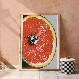 May include: A framed wall art print featuring a stylized, graphic illustration of a sliced grapefruit. The citrus fruit displays vibrant orange-red segments with a yellow rind, set against a light gray background with a subtle dot-matrix texture.