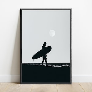 May include: A framed wall art print featuring a black silhouette of a person carrying a surfboard along a shoreline at night. The scene includes a dark ocean, a sandy beach, and a bright full moon set against a light gray sky in a vertical orientation.