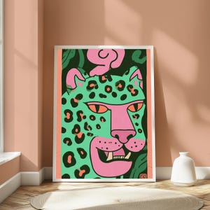 May include: A framed art print featuring a stylized illustration of a leopard head. The leopard is mint green with black spots, a pink snout, and orange eyes. The background is dark green with pink abstract shapes, all enclosed in a pink border.