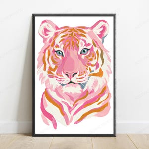 May include: A framed wall art print featuring a stylized tiger head illustration. The tiger is rendered in shades of pink, white, and orange with light blue eyes. The artwork is enclosed in a thin black frame and displayed against a white wall.
