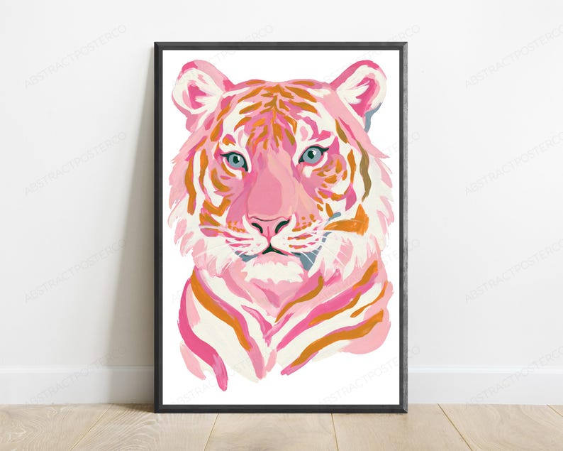 May include: A framed wall art print featuring a stylized tiger head illustration. The tiger is rendered in shades of pink, white, and orange with light blue eyes. The artwork is enclosed in a thin black frame and displayed against a white wall.