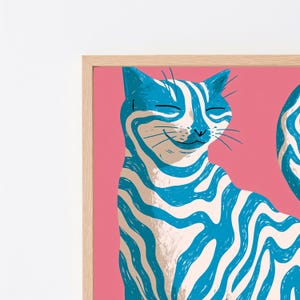 May include: A framed wall art print featuring a stylised cat with blue and white wavy stripes against a solid pink background. The cat has closed eyes and a subtle smile, rendered in a graphic illustration style within a light wood frame.