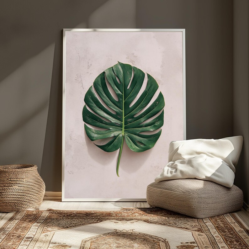 May include: A framed wall art print featuring a single, large, deep green monstera leaf centered against a soft, pale pink textured background. The artwork is enclosed in a thin white frame, showcasing the detailed veins and lobes of the leaf.